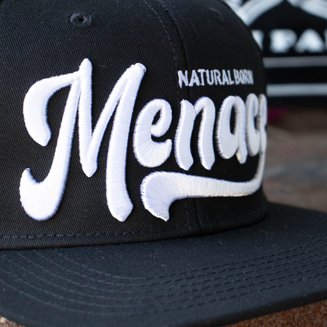 Close up of front of Menace Clothing black baseball hat with white puff embroidered Natural Born Menace. 